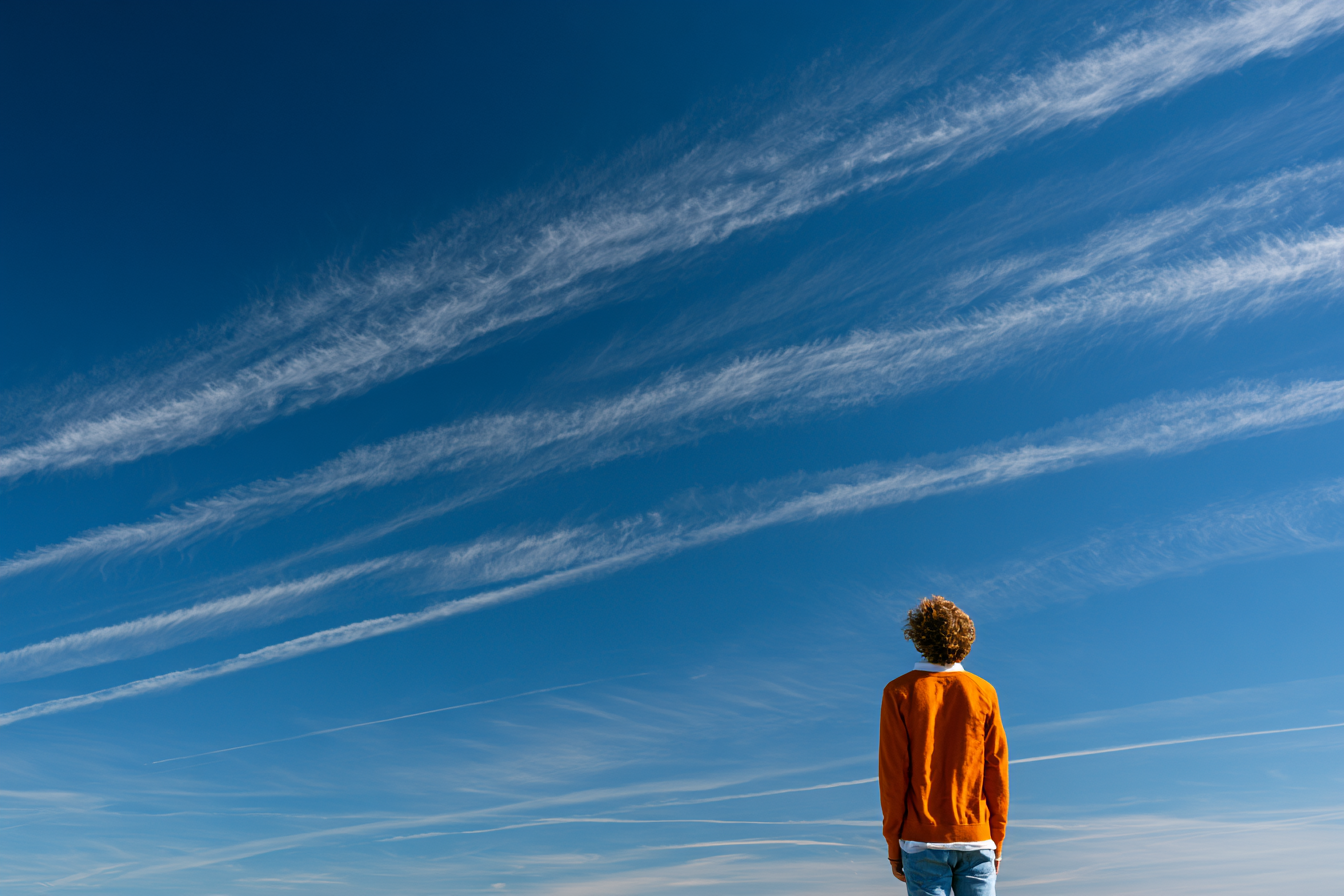 A lone young person stands beneath a vast open sky crossed by a few faint jet contrails.